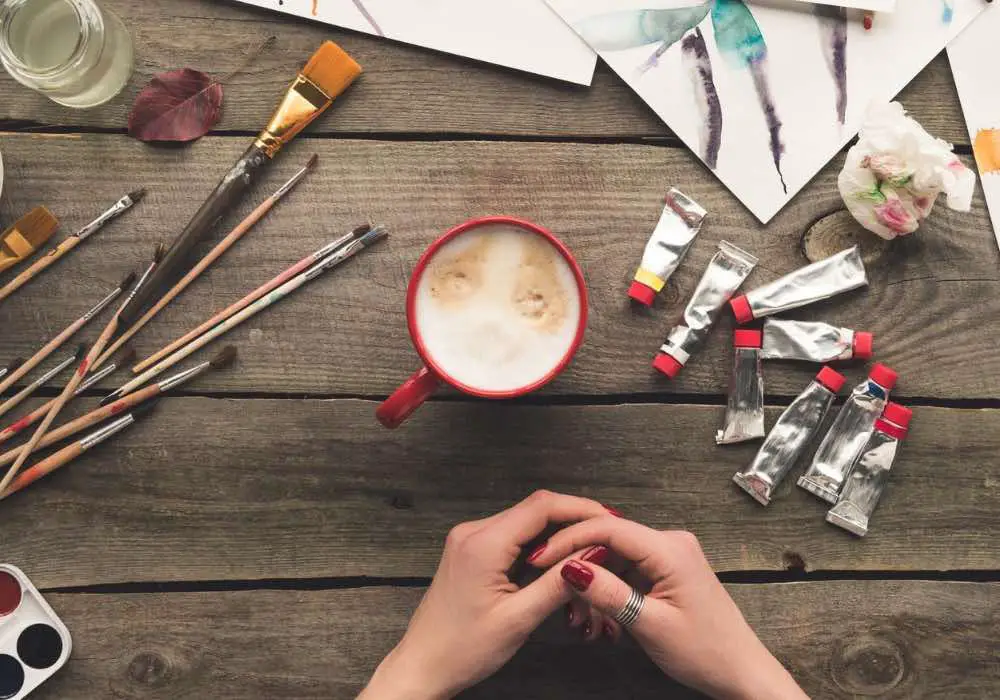 A cup of coffee surrounded by paint tubes, brushes and paper